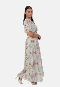 Robe maxi florale à manches courtes, avec des motifs beiges, marron et bleus sur un fond crème. Fabriquée en tissu léger.