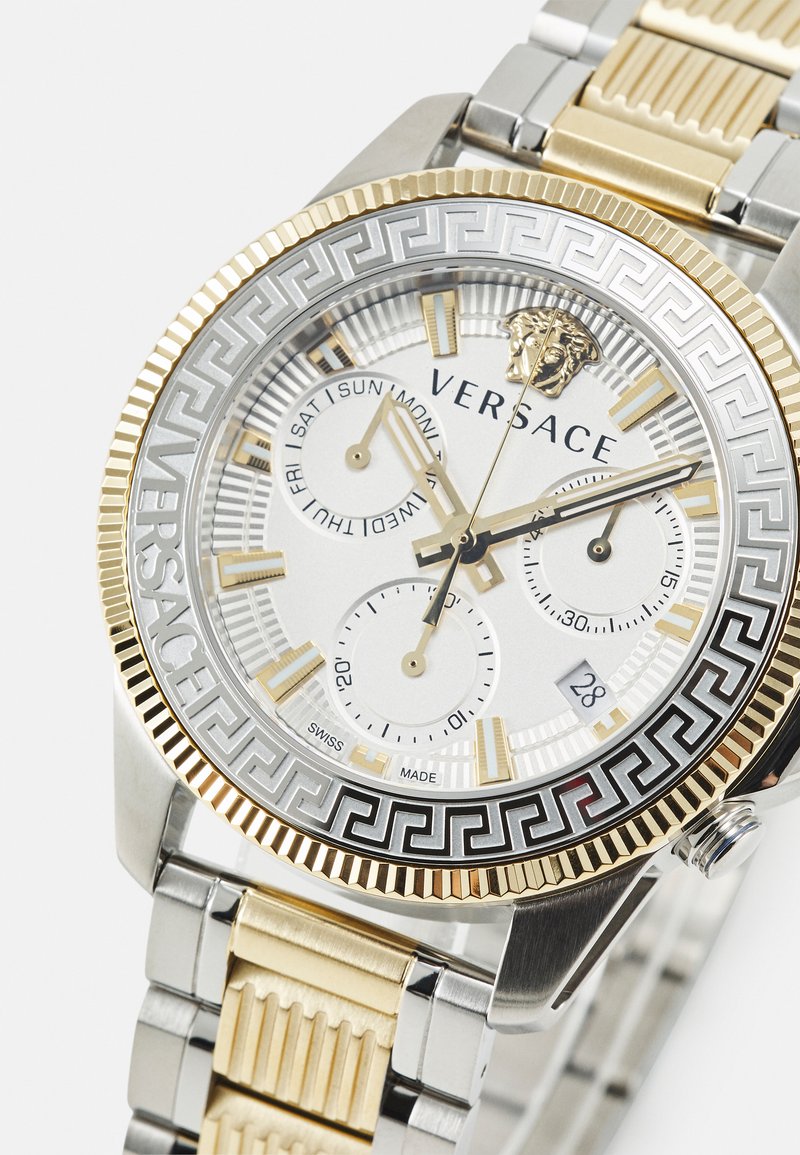 Montre Versace avec un bracelet en acier inoxydable argent et or. Le cadran rond argenté présente des accents dorés, trois sous-cadrans et un motif grec complexe.