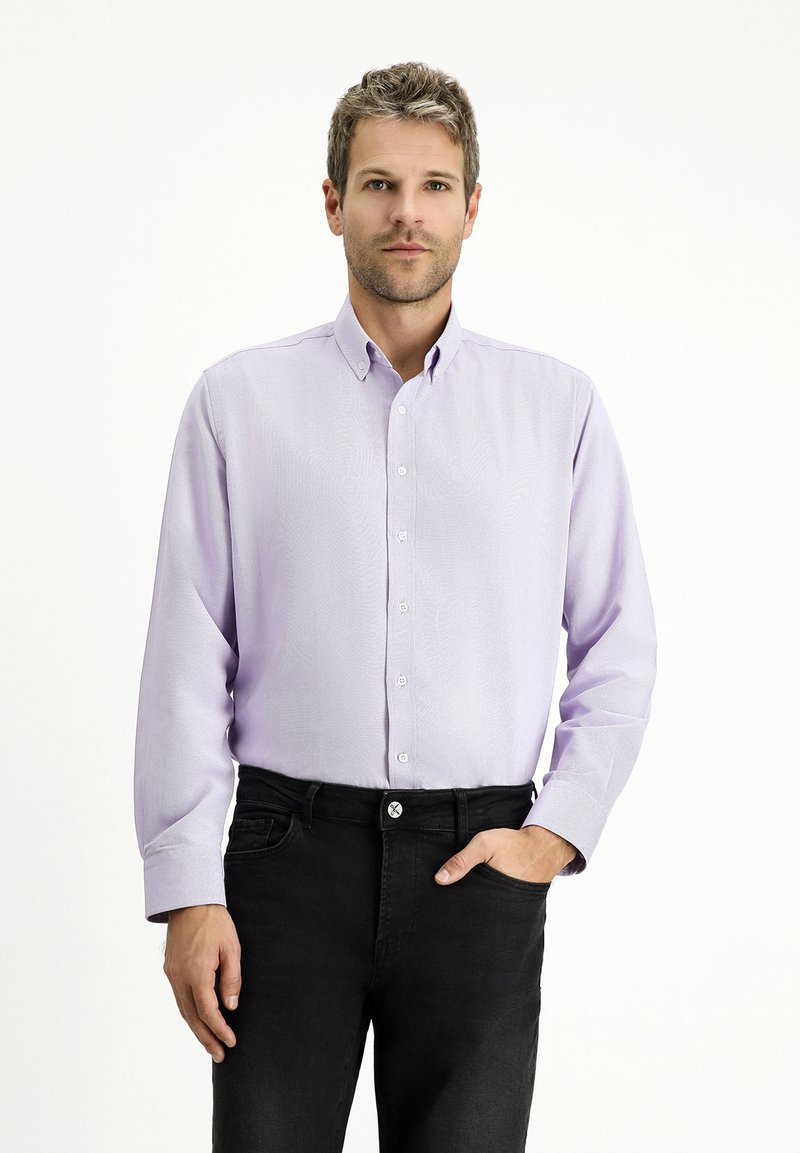 Jasnofioletowa koszula na guziki z teksturowanym wzorem i kołnierzykiem, połączona z czarnymi dopasowanymi dżinsami. Model stoi z jedną ręką w kieszeni.