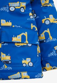 Jachetă impermeabilă albastră cu un model de vehicule de construcție galbene, inclusiv camioane basculante și excavatoare, având un detaliu al buzunarului din față.