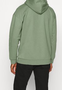 Persona che indossa una felpa con cappuccio verde chiaro e pantaloni neri, vista di spalle su uno sfondo neutro.