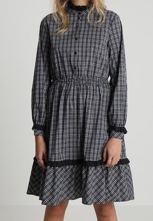 Frau trägt ein langärmeliges schwarz-weiß kariertes Kleid mit gerafftem Kragen, Manschetten und Saum, steht vor einem schlichten Hintergrund.