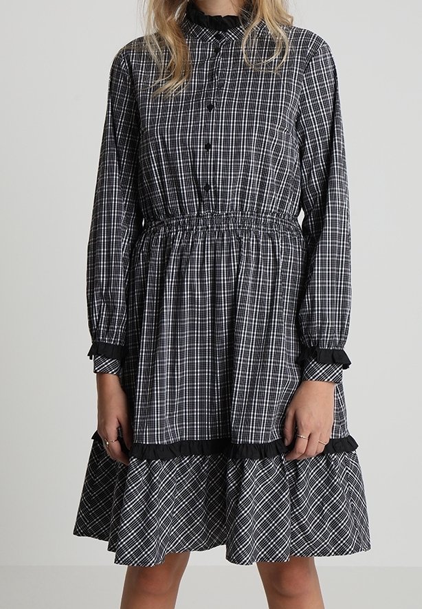 Femme portant une robe à manches longues à carreaux noir et blanc avec un col, des poignets et un ourlet à volants, debout devant un fond uni.