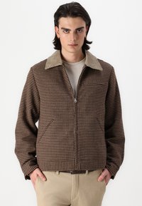 Chaqueta marrón a cuadros con cuello de pana, cierre de cremallera y bolsillos laterales. Llevada sobre una camisa beige, combinada con pantalones de color claro.