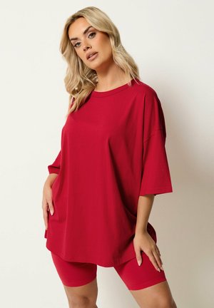 Blonde Frau posiert in übergroßem rotem T-Shirt und passenden roten Radlerhosen vor schlichtem hellem Hintergrund.