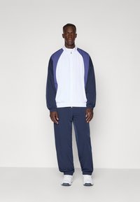 Trenirka z belimi in modrimi barvnimi bloki, z jakno na zadrgo in hlačami z elastičnimi manšetami, prikazana na modelu.