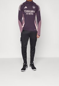Bordeauxfarbene Langarm-Trainingsjacke mit halbem Reißverschluss und weißen Akzenten. Schwarze Hosen mit seitlichen Streifen. Das Model trägt schwarze Sportschuhe.