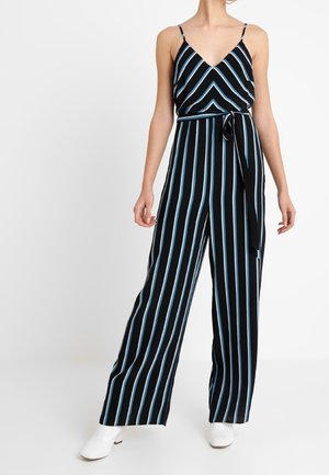 Femme portant une combinaison noire avec des rayures verticales bleues et blanches, une ceinture à nouer, des bretelles spaghetti et des bottines blanches.