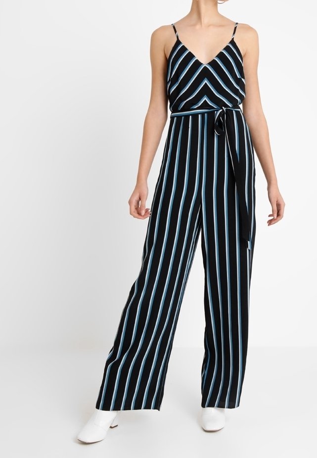 Femme portant une combinaison noire avec des rayures verticales bleues et blanches, une ceinture à nouer, des bretelles spaghetti et des bottines blanches.