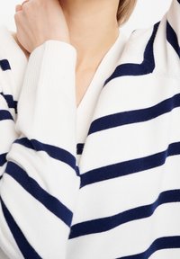 Valkoinen neulepusero, jossa on tummansinisiä vaakaraitoja, V-pääntie ja ribbireunukset. Pehmeä rakenne, jossa näkyvä neulekuvio. Lähikuva.