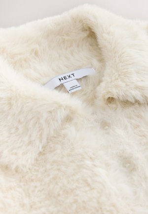 Kermanvärinen tekoturkisvaate, jossa on kaulus, nappikiinnitys ja "NEXT"-merkillä varustettu lappu. Pehmeä tekstuuri ja pörröinen ulkonäkö.