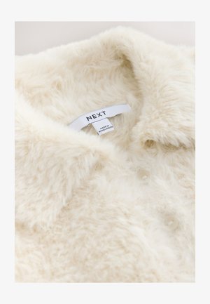 Kermanvärinen tekoturkisvaate, jossa on kaulus, nappikiinnitys ja "NEXT"-merkillä varustettu lappu. Pehmeä tekstuuri ja pörröinen ulkonäkö.