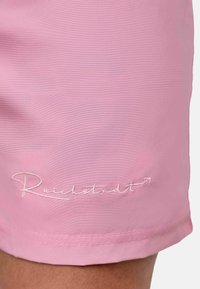 Pantaloni shorts rosa realizzati in tessuto liscio con un logo bianco ricamato che dice "Reichetstadt" vicino all'orlo. Design semplice con linee pulite.