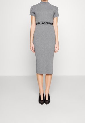 Kobieta nosząca dopasowaną szarą prążkowaną sukienkę midi z krótkimi rękawami i napisem „Karl Lagerfeld” na talii, zestawioną z czarnymi, szpiczastymi szpilkami.
