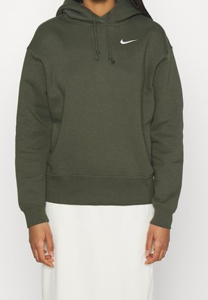 Osoba ubrana w ciemnozieloną bluzę z kapturem Nike z sznureczkami i białym logo Swoosh, w połączeniu z białą spódnicą, ręce swobodnie opuszczone wzdłuż ciała.