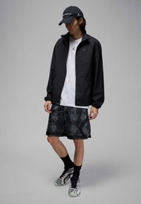 Chaqueta negra con cremallera, camiseta blanca, pantalones cortos negros con estampado, y zapatillas grises con acentos verdes. Los accesorios incluyen una gorra negra.
