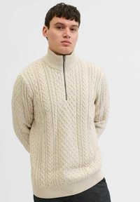 Beżowy sweter z zamkiem, z wzorem w warkocze, z wysokim kołnierzem i ściągaczami przy mankietach oraz dole. Miękka tekstura, gruby splot. Prosty design.