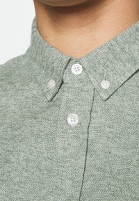 Zelená košile s texturou a límečkem na knoflíky, se dvěma bílými knoflíky a jemným tkaným vzorem. Detailní pohled na výstřih.