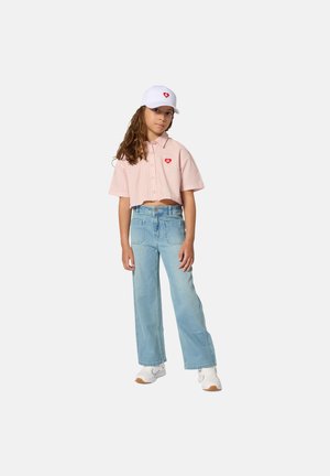 Chemise raccourcie rayée rose à manches courtes avec un patch en forme de cœur rouge, portée avec un jean large bleu clair et une casquette blanche. Baskets blanches.