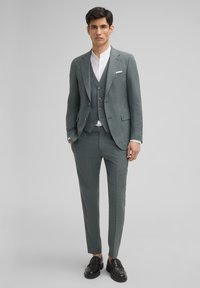 Grau strukturierter Anzug mit schmal geschnittenen Hosen, Weste und Blazer. Darunter ein weißes Hemd. Schwarze Slipper verleihen dem Outfit einen eleganten Touch.
