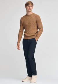 Brauner Pullover mit Zopfmuster und rundem Ausschnitt, kombiniert mit dunkler Hose und hellen Sneakers, mit kleinem Logo auf der Brust.