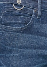 Dark blue denim jeans featuring subtle whiskering, orange stitching, and a metal button with logo detail on the waistband.