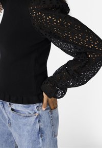 Musta paita, jossa on teksturoituja puffihelmoja ja leikkauksia, yhdistettynä vaaleansinisiin farkkuihin. Paidassa on frilloreunus.