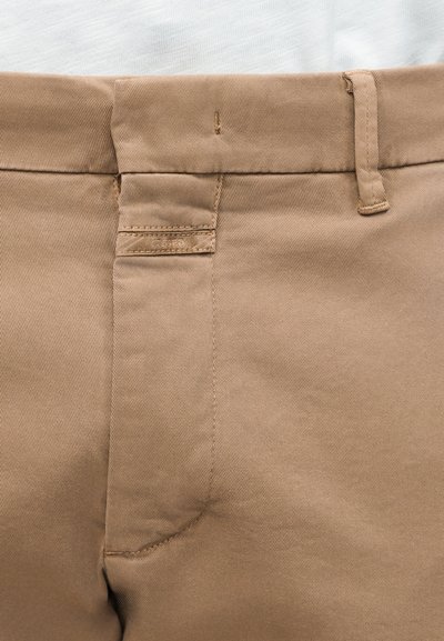 Gros plan sur un pantalon beige à l'avant avec passants de ceinture, fermeture éclair et petite étiquette cousue sur la ceinture.