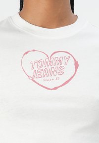 Balts kokvilnas T-krekls ar plašu apaļu kakla izgriezumu, kurā attēlots rozā sirdis grafiks ar uzrakstu "TOMMY JEANS Since 85" treknā fontā.