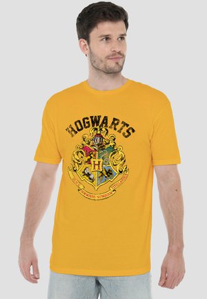 Mann trägt ein gelbes T-Shirt mit einem abgenutzten Hogwarts-Wappen und lateinischem Motto und steht vor einem schlichten hellen Hintergrund.