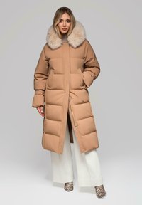 Lungo cappotto imbottito di colore cammello con colletto in faux fur, caratterizzato da una linea dritta, bottoni a scatto e tasche laterali. Indossato sopra pantaloni bianchi larghi.