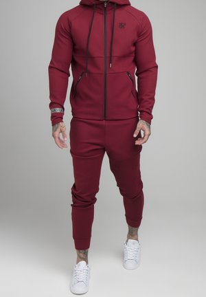 Homme portant un sweat à capuche zippé bordeaux et un pantalon de jogging assorti, associé à des baskets blanches, debout devant un fond gris uni.