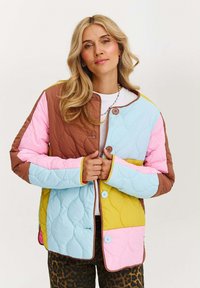 Žena s dlhými blond vlasmi, oblečená v mnohobarevnej prešívanej bunde a nohaviciach s leopardím vzorom, stojí na pozadí s bielou farbou.