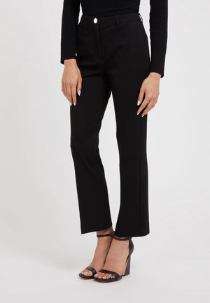 Zwarte op maat gemaakte pantalons met rechte pijpen, voorzien van een tailleband met een knoopsluiting en zijzakken; gecombineerd met zwarte haksandalen.