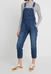 Blå denimoveraller med justerbara remmar, kortade ben, framfickor, bärs med en svart och vit randig polotröja och orangea sneakers.