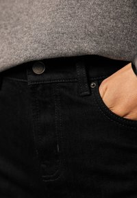 Sorte jeans i denim med en tekstureret grå striktrøje, der overlapper taljebåndet. Har en metal knap og en hånd hvilende i lommen.