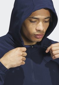 Granatowa kurtka z kapturem, z ćwierć zamkiem, teksturowana tkanina, elastyczne mankiety i sznurek przy kapturze. Dopasowany krój wokół twarzy.