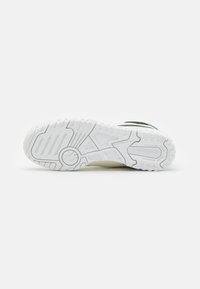 Semelle de sneaker blanche New Balance orientée vers le haut, montrant des motifs de grip texturés et le nom de la marque sur un fond blanc lisse.