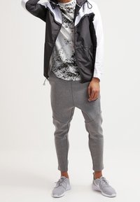 Svart och vit vindjacka, grå sweatpants, skjorta med splatter-mönster och grå sportskor. Lätta material, åtsittande design.