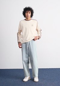 Μπεζ πουκάμισο μακρυμάνικο με καφέ λεπτομέρειες και λογότυπο, συνδυασμένο με ανοιχτό μπλε φαρδύ τζιν και λευκά αθλητικά παπούτσια σε φόντο μπλε χαλιού.