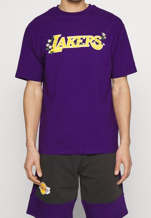 Camiseta corta de color púrpura con mangas cortas que presenta "LAKERS" en amarillo con acentos florales. Combinada con pantalones cortos grises, que muestran el logo del equipo en la pierna.