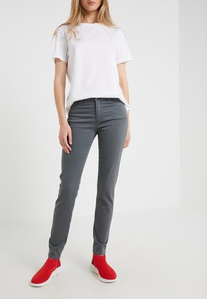 Kobieta ubrana w biały krótki rękaw, szare dopasowane spodnie oraz czerwone wsuwane trampki, stojąca na tle jednolitej białej ściany.