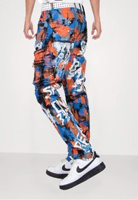 Cargo-Hosen mit einem mehrfarbigen Spritzmuster in Blau, Orange und Schwarz. Mit Reißverschlüssen, Taschen und weißem Schriftzug. Weißer Gürtel und Sneakers.