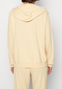 Béžová mikina s kapucí vyrobená z měkké látky, s uvolněným střihem a pružnými manžetami, s hladkým povrchem a bez viditelných vzorů.