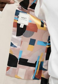 Veste beige sur mesure avec une doublure colorée et à motifs présentant des formes géométriques en rose, bleu, jaune et noir. Étiquette visible.