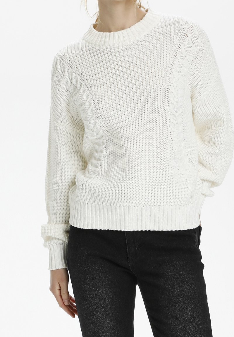 Biały sweter dziany z teksturowanym wzorem oraz ściągaczami na mankietach i dole. Połączony z czarnymi jeansami o luźnym kroju.