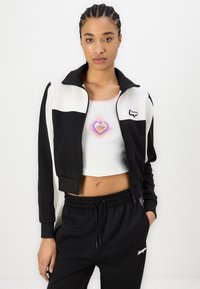 Veste courte zippée noir et blanc avec un col montant, associée à un crop top blanc avec un motif en forme de cœur, et un jogging noir à taille élastique.