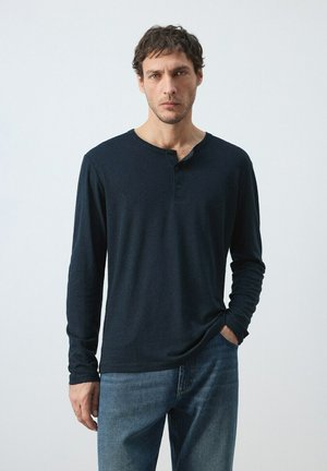 Hombre de pie con la mano en el bolsillo, vistiendo una camiseta henley de manga larga azul marino y vaqueros azules contra un fondo liso y claro.