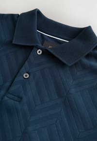 Cămașă polo bleumarin cu un model geometric texturat, având o guler tricotat, două butoane negre și o dublură albă la interiorul gulerului.