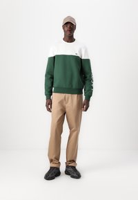 Sudadera color bloqueada en verde y blanco con puños y dobladillo acanalados, pantalones beige a juego y zapatillas negras. Una gorra beige completa el look.